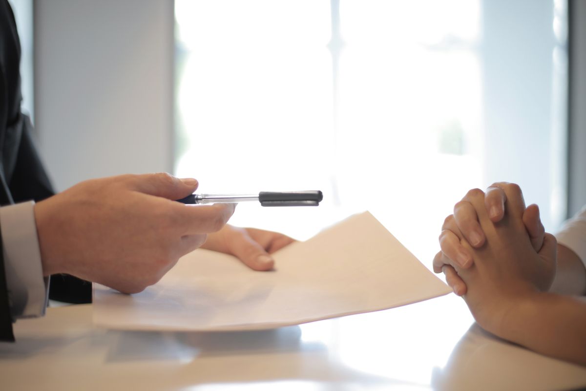 Two professionals seated across a white table, one presenting a document with a pen, in a brightly lit office